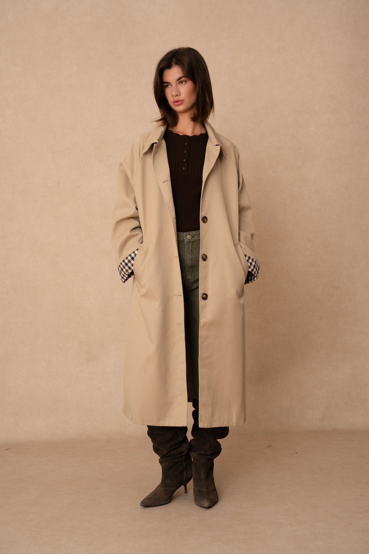 Model wearing a long taupe trench coat with a button front, collar and check-pattern turned-back cuffs.