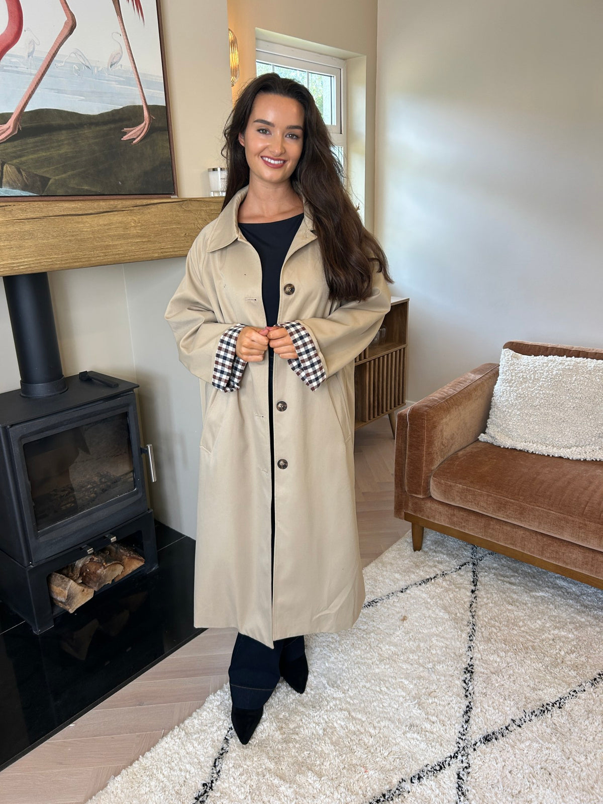 Model wearing a taupe trench coat with a button front, long sleeves and turned-back check cuffs.